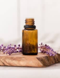 Amber colored essential oil bottle on wooden cutting board with lavender sprigs behind it.