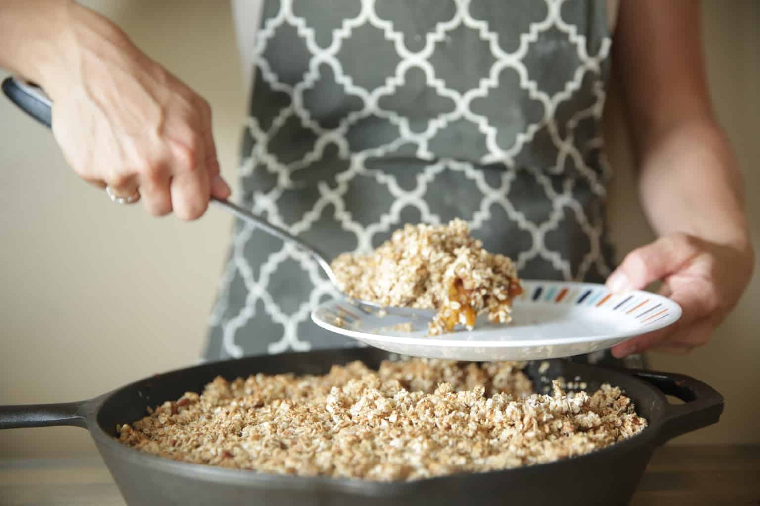 scooping homemade peach crisp from cast-iron skillet onto small plate