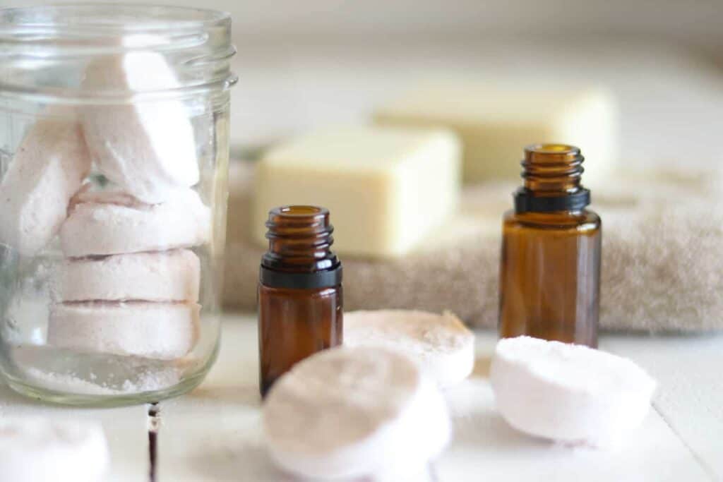 shower melt tablets with soap bars and glass essential oil bottles on white table.