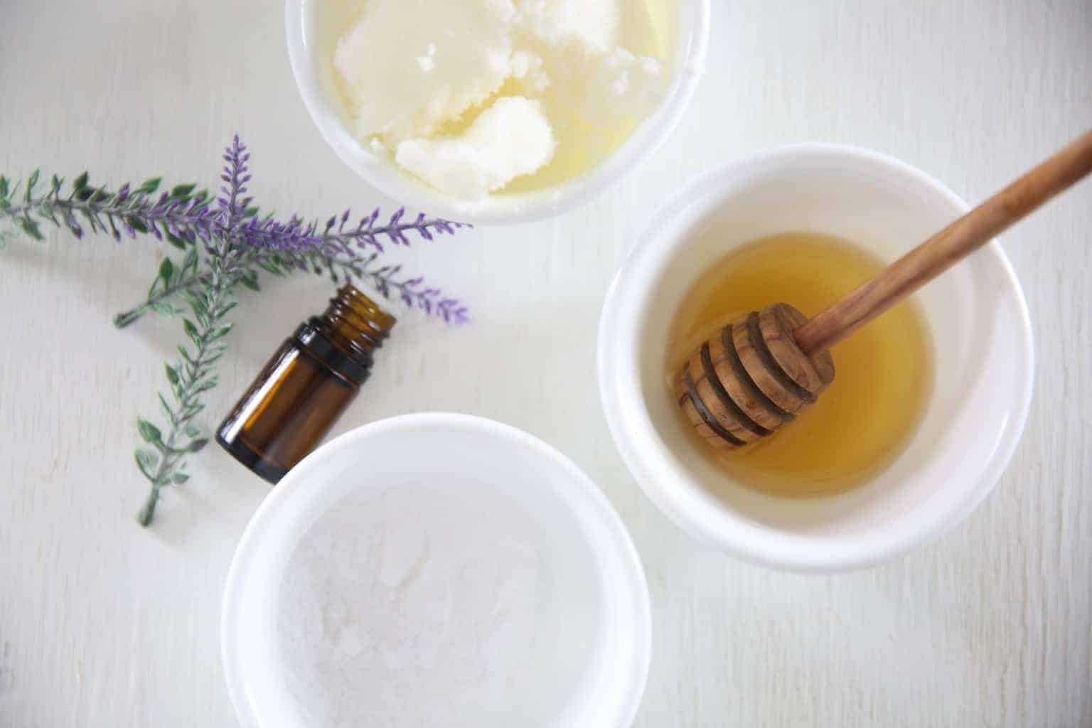 Beef tallow and honey in separate white bowls on white table with essential oil bottle and lavender leaves.