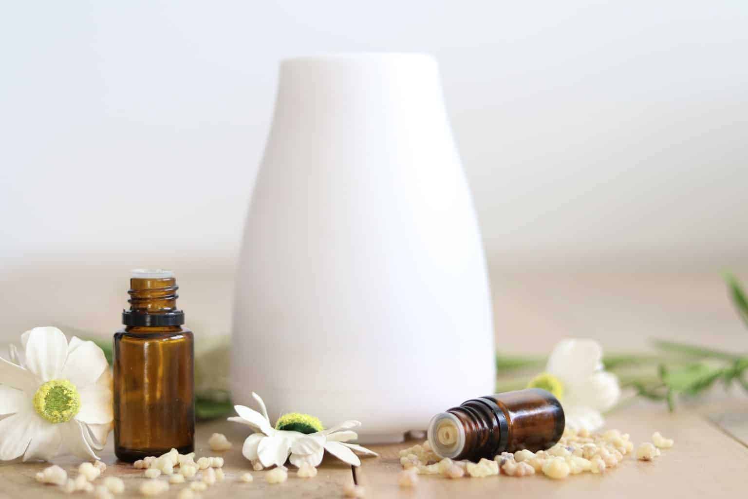 White essential oil diffuser surrounded by flowers and oil bottles on wooden table.