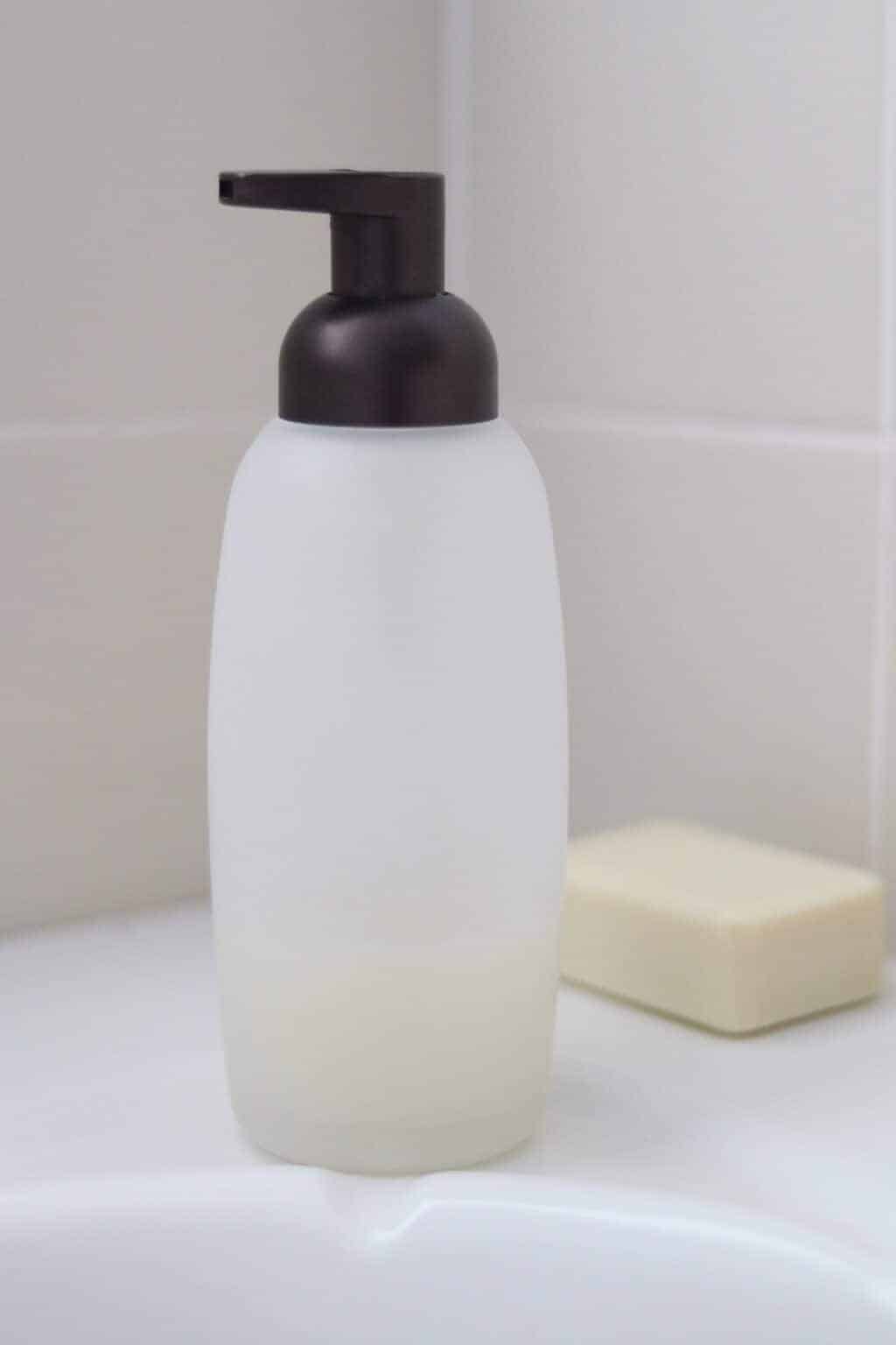 Glass shampoo container with soap bar on bathtub edge.