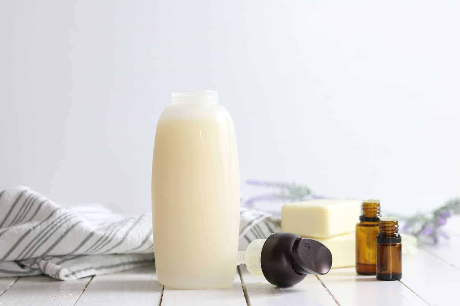 Glass dispenser of homemade shampoo, soap bars, oil bottles and decorative towel on white shiplap.