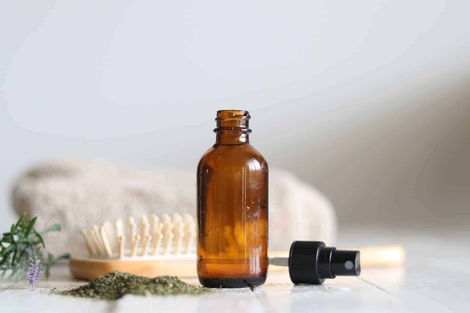 Spray bottle of homemade hair serum with hair brush and herbs in background.