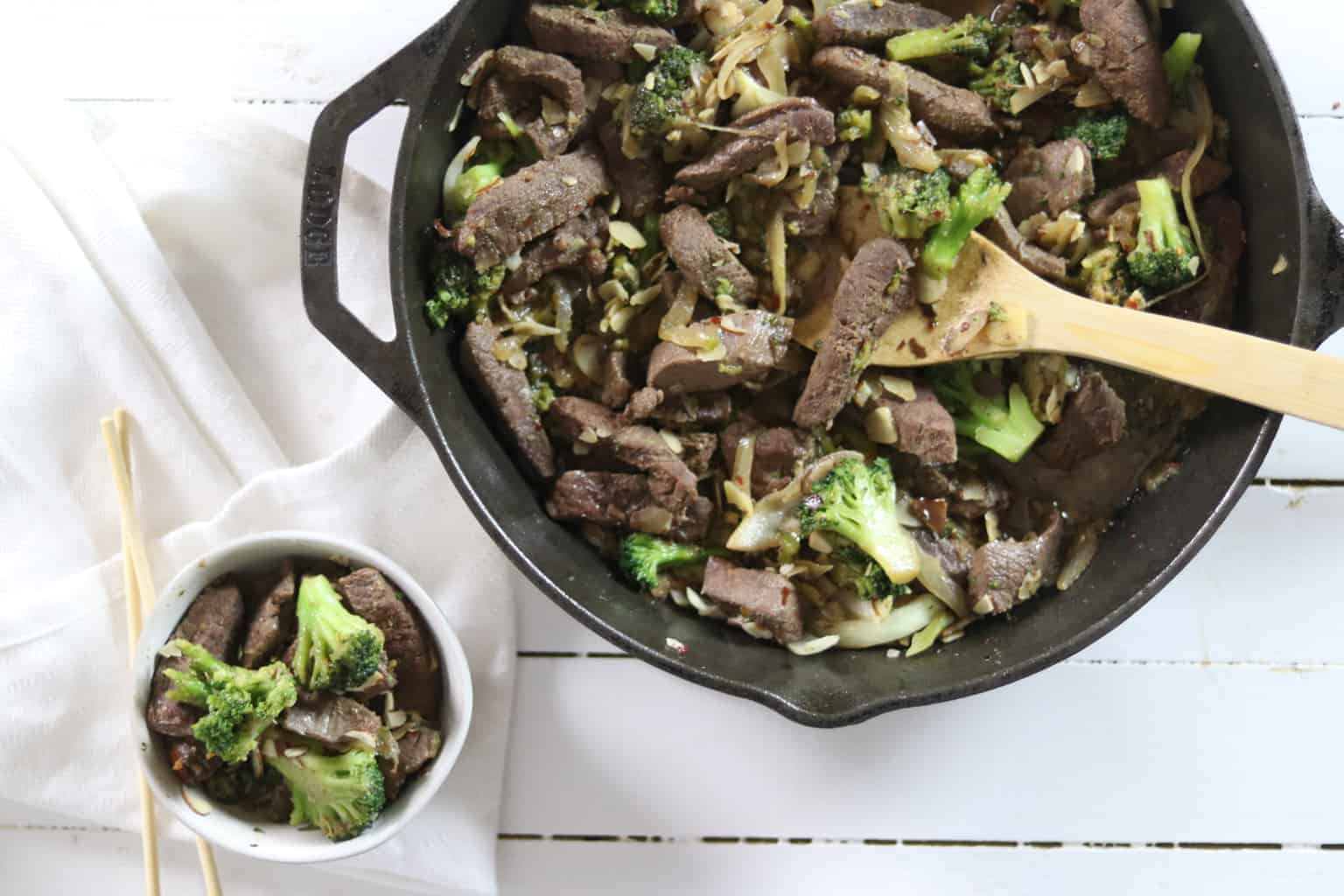 Cast iron skillet with broccoli and beef on white wooden table.