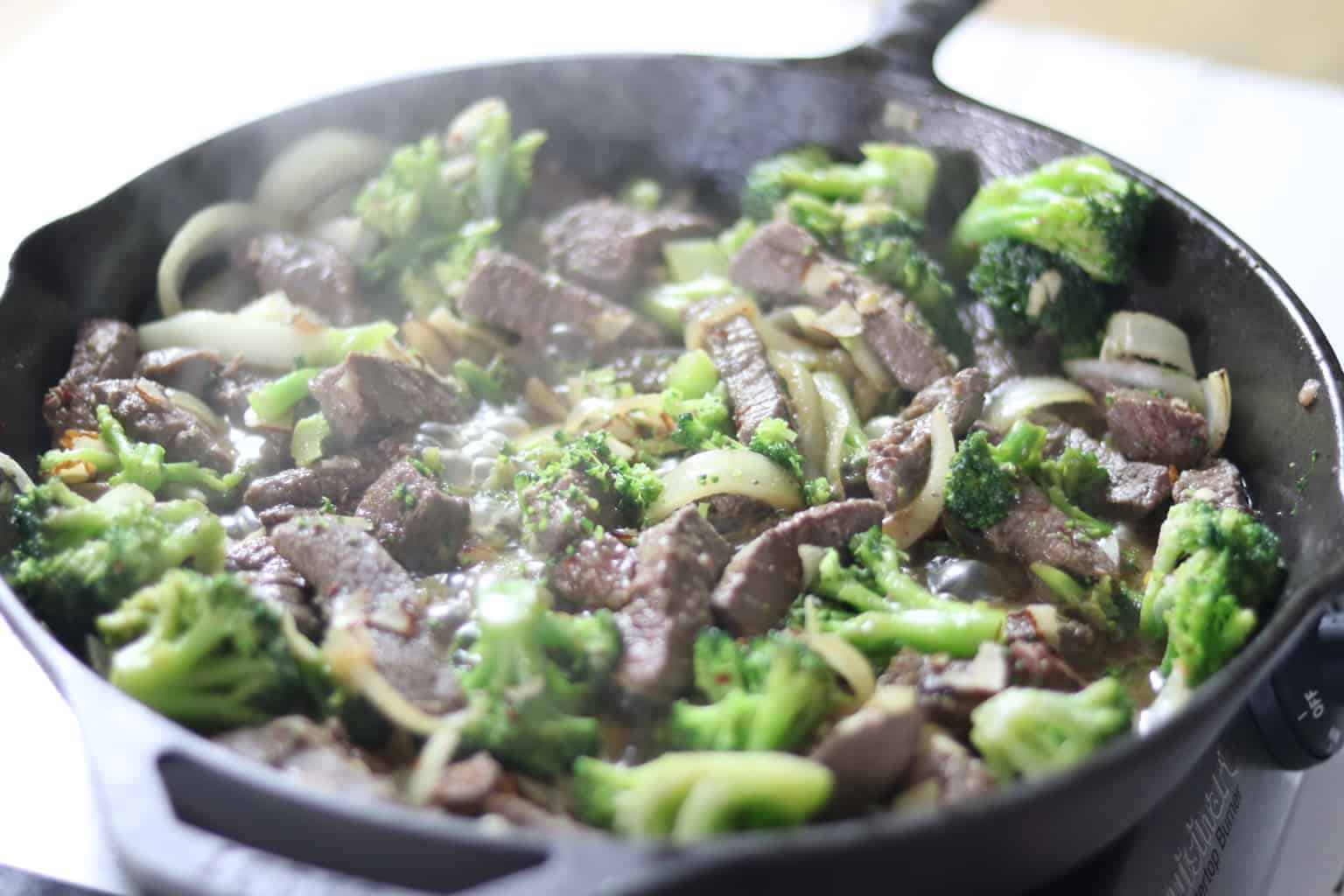 Broccoli, beef, and onions cooking in cast iron skillet. 