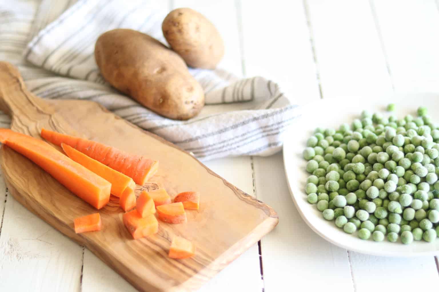 Chopped carrots on wooden cutting board, frozen peas on white plate, and potatoes.
