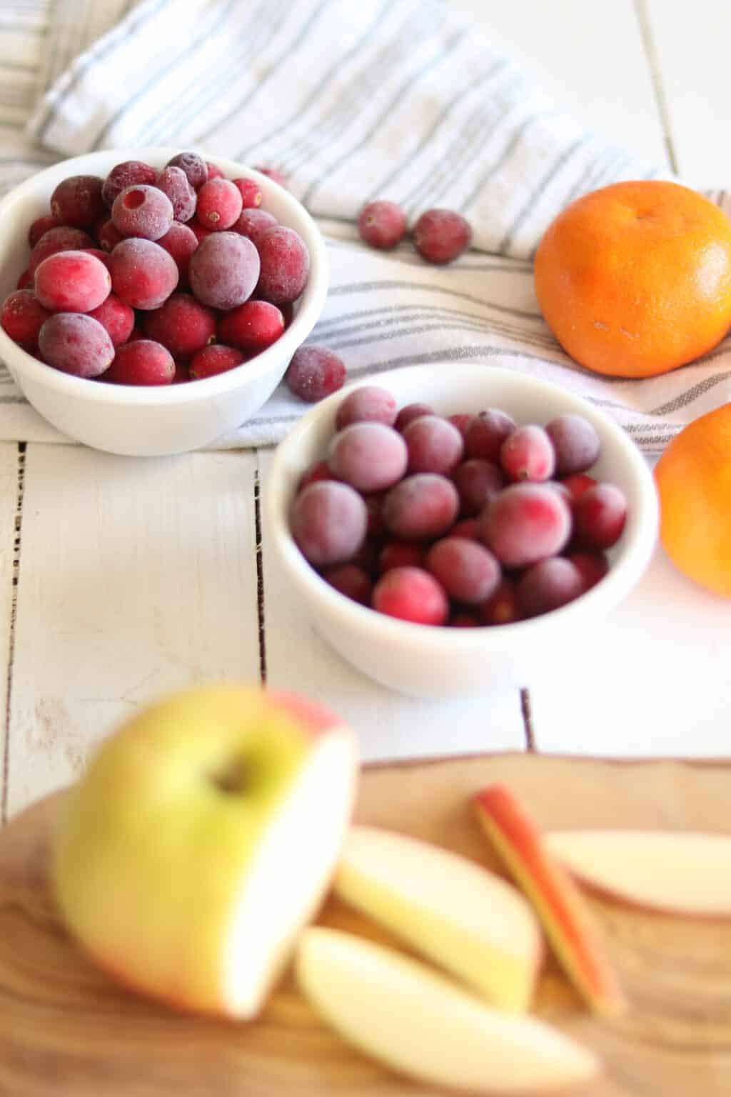 Frozen cranberries in white bowl, sliced apple, and fresh clementine's on table.