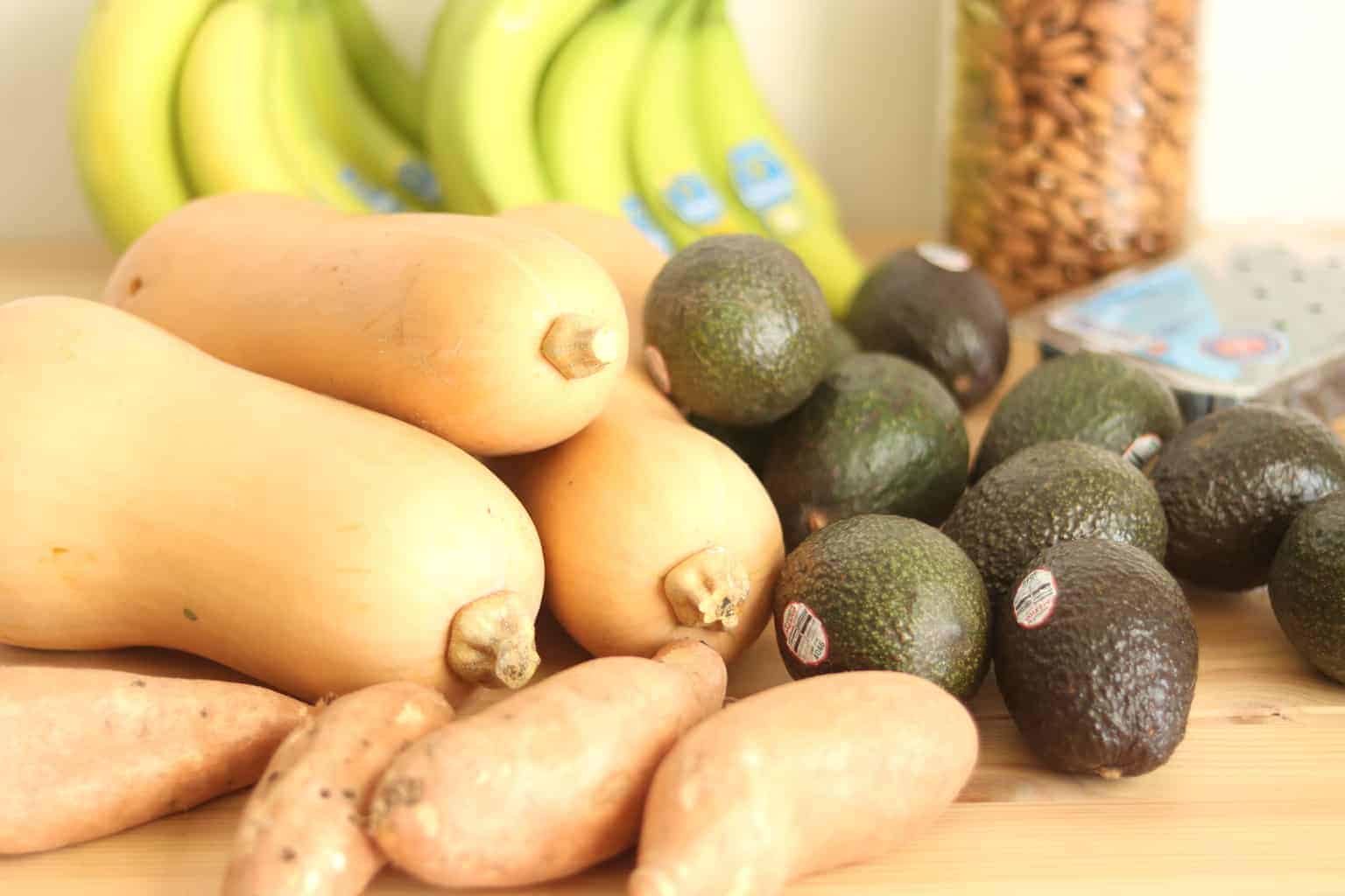 Squash, sweet potatoes, avocado, and bananas on wooden table.