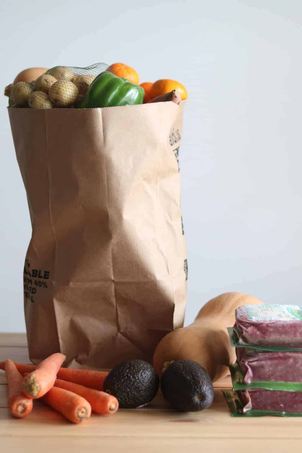 Fresh veggies and meet in brown paper shopping bag and on table.