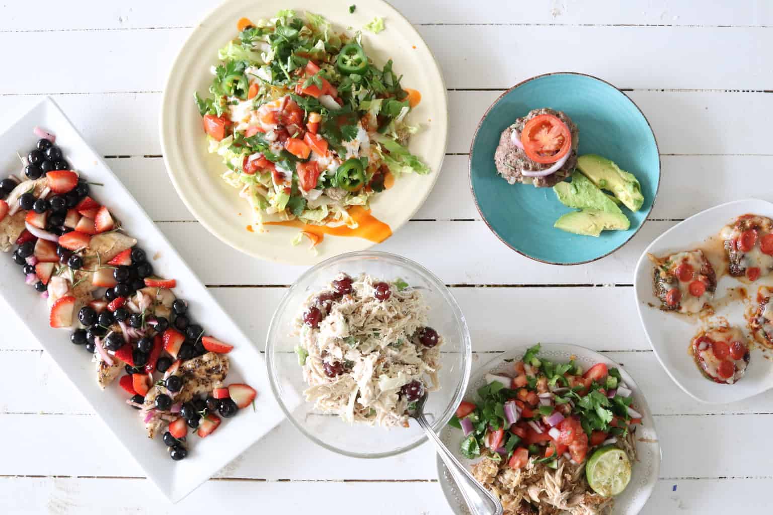 Salad, chicken salad, and zucchini pizzas in dishes on wooden table.