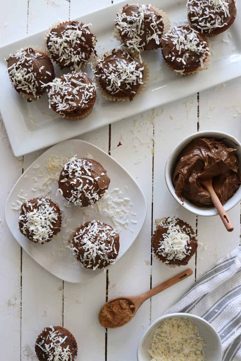 Vanilla cupcakes with chocolate icing topped with coconut on white plates.