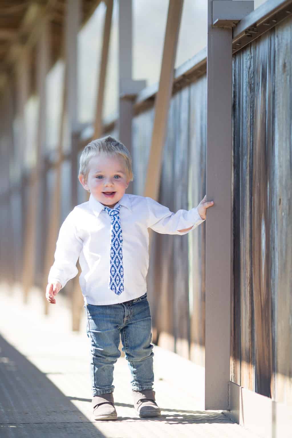 Picture of toddler in white button down and blue tie standing on wood covered bridge.