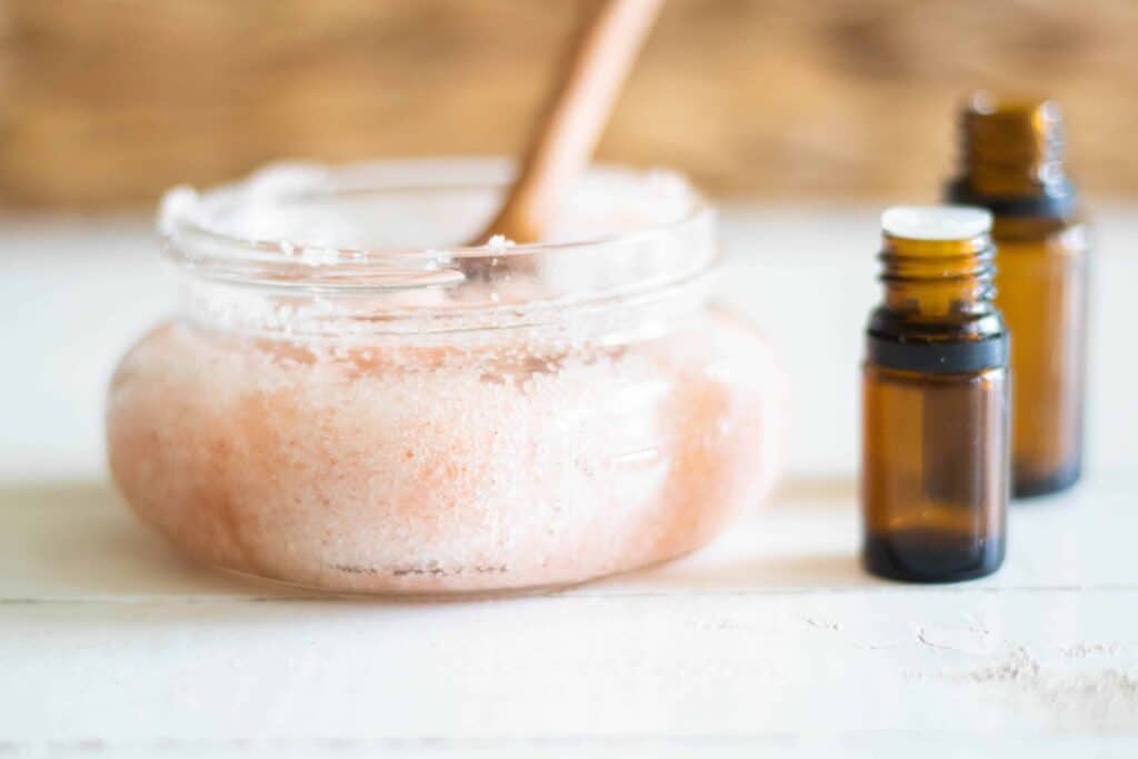 Pink foot scrub in mason jar with wooden spoon in it and amber essential oil bottles on white table.