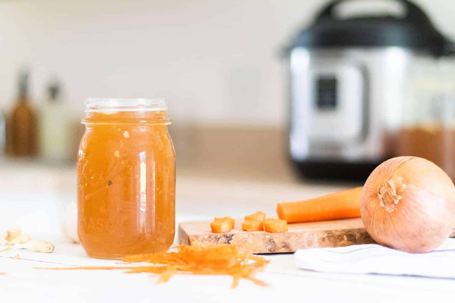 Bone broth in mason jar with fresh carrots and onion on counter with instant pot in background.