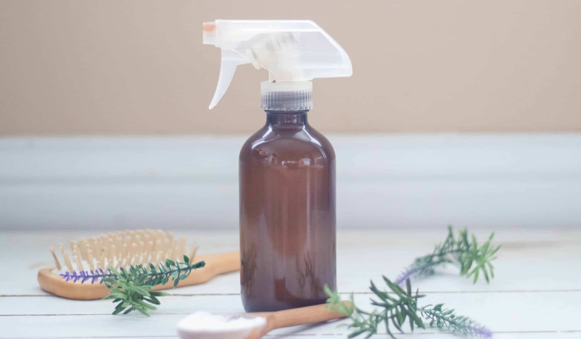 Medium sized amber color glass dry shampoo spray bottle sitting on white ship lap with rosemary and lavender sprigs around.