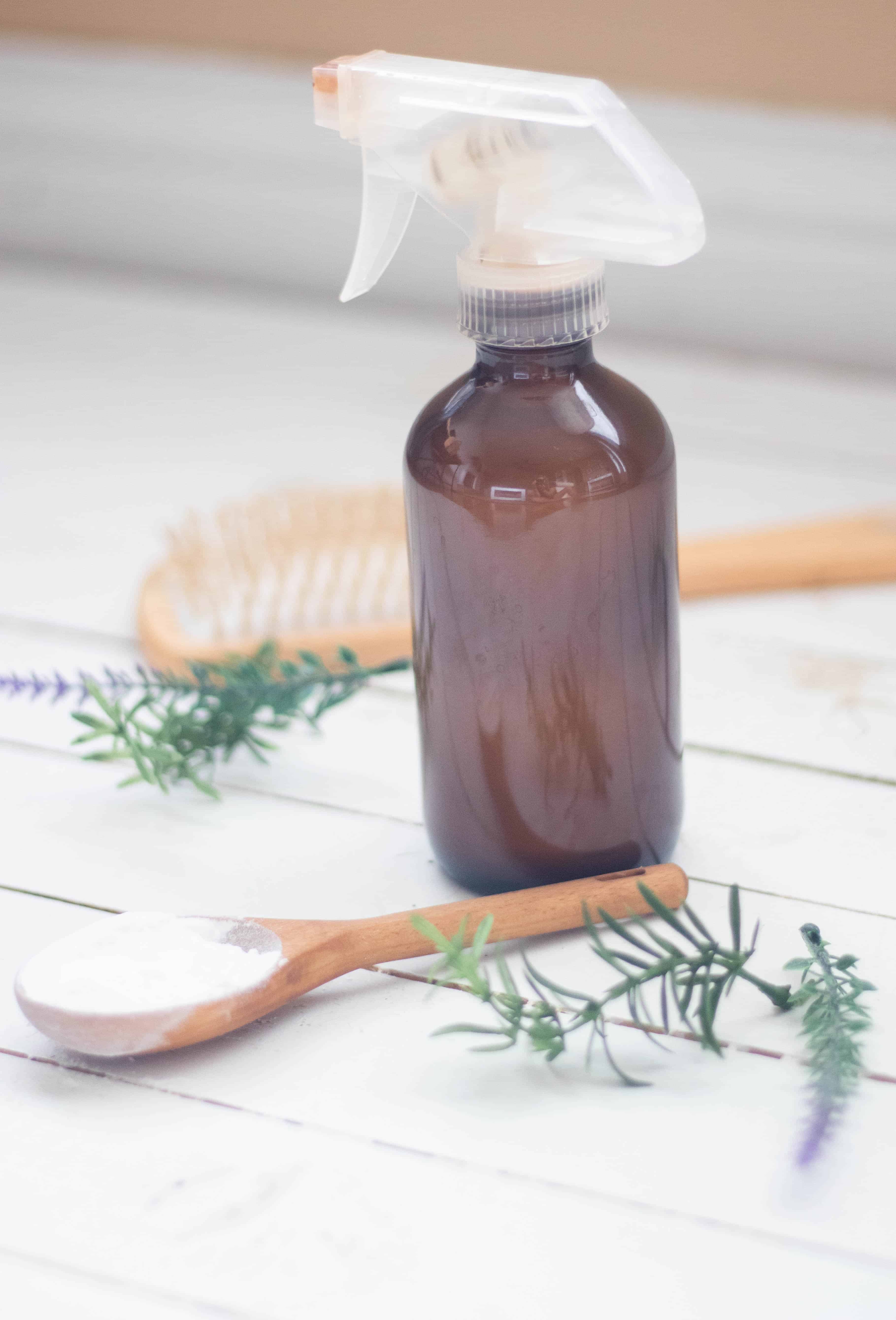 Amber colored spray bottle, wooden measuring spoon, and tan brush laying on white ship lap.