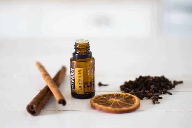 Tangerine essential oil bottle, cinnamon sticks, orange slice and clove buds on table.