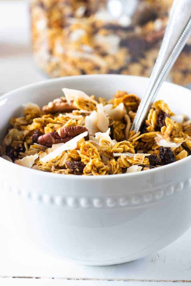 Homemade pumpkin granola in white bowl with nuts and coconut chips on top.