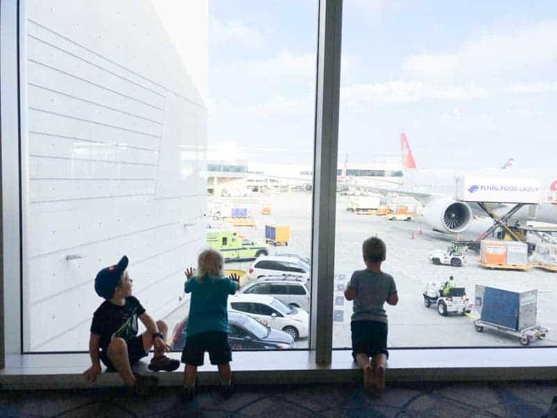 Three boys looking out the window at an airport apron before their flight.