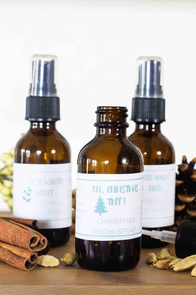 Glass spray bottles with Christmas labels on wooden table surrounded by cinnamon sticks and pine cones.