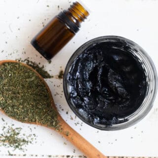Homemade black drawing salve in small mason jar with essential oil bottle and dried herbs on a wooden table.