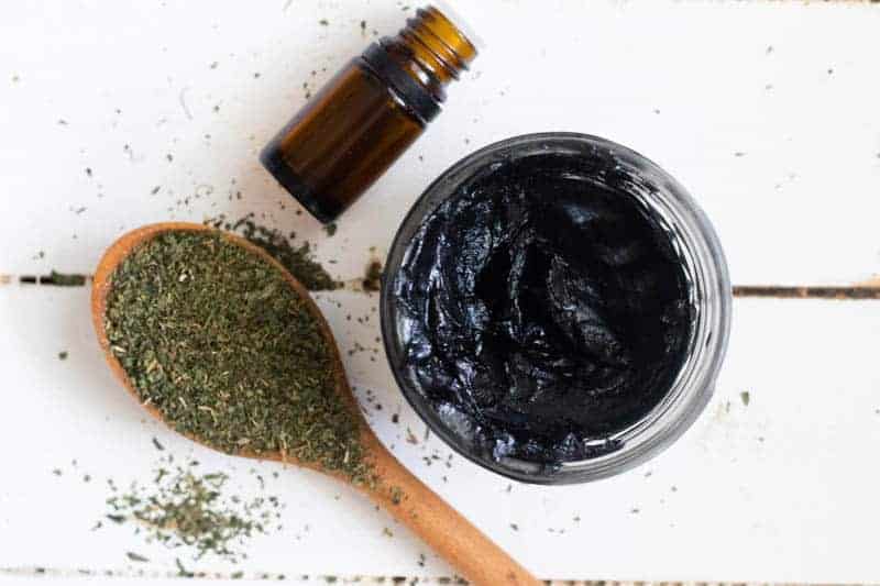 Homemade black drawing salve in small mason jar with essential oil bottle and dried herbs on a wooden table.