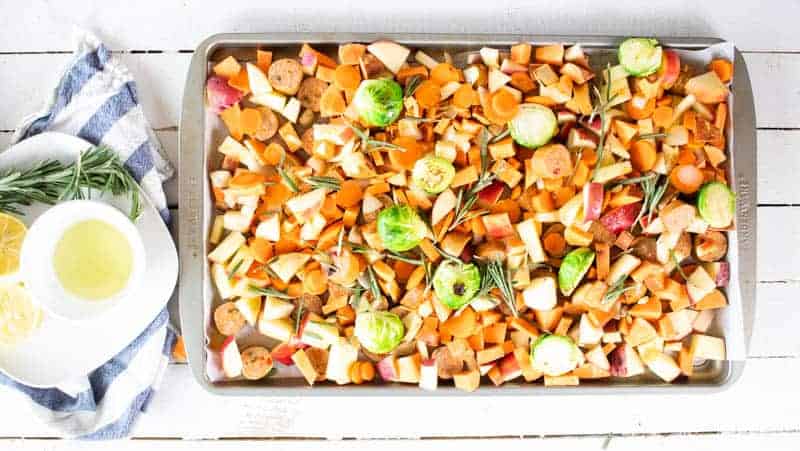 Sausage, apple, and vegetables on sheet pan on white wooden table.