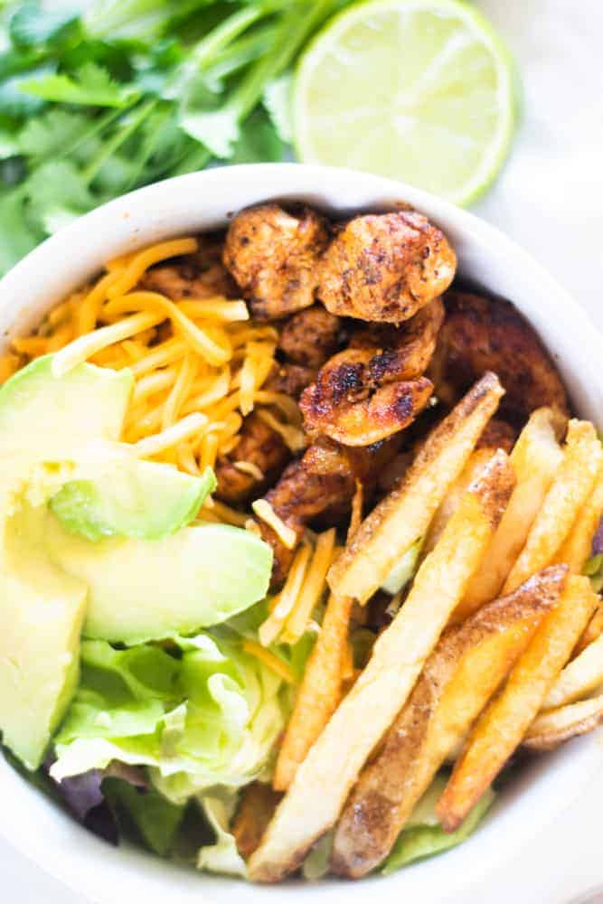 Fresh lettuce in white bowl with chicken, avocado, cheese and French fries.