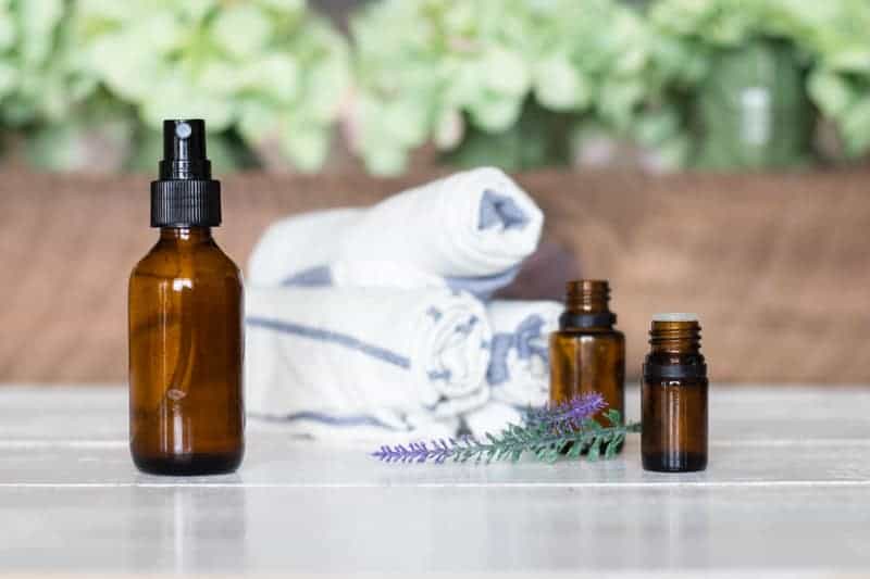 Glass spray bottle with essential oil bottles, lavender sprigs and towels on wood table.