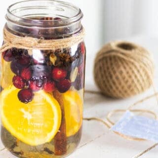 Cranberries orange slices fresh herbs and cinnamon sticks in mason jar with water for stovetop potpourri.