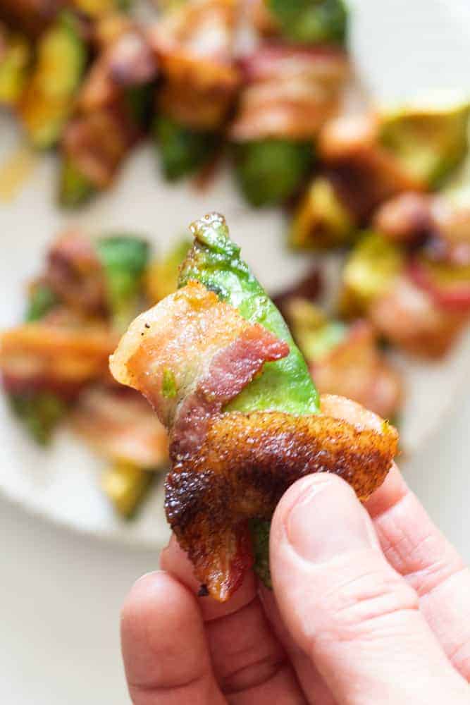 Hand holding bacon wrapped avocado fry with plate of bacon wrapped avocado fries in background.