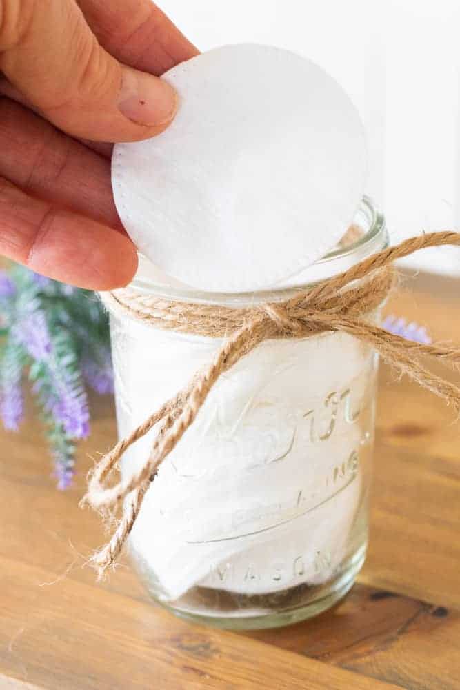 Hand holding white cotton round over small mason jar of makeup remover.