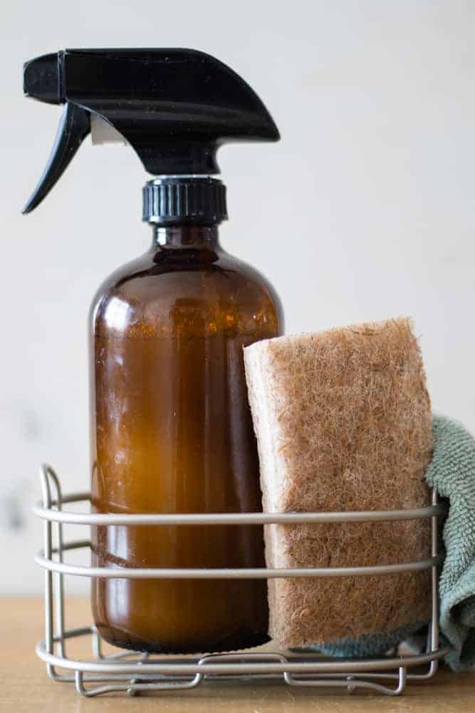 DIY granite countertop cleaner in an amber spray bottle on a metal rack with a scrub brush and micro-fiber towel.