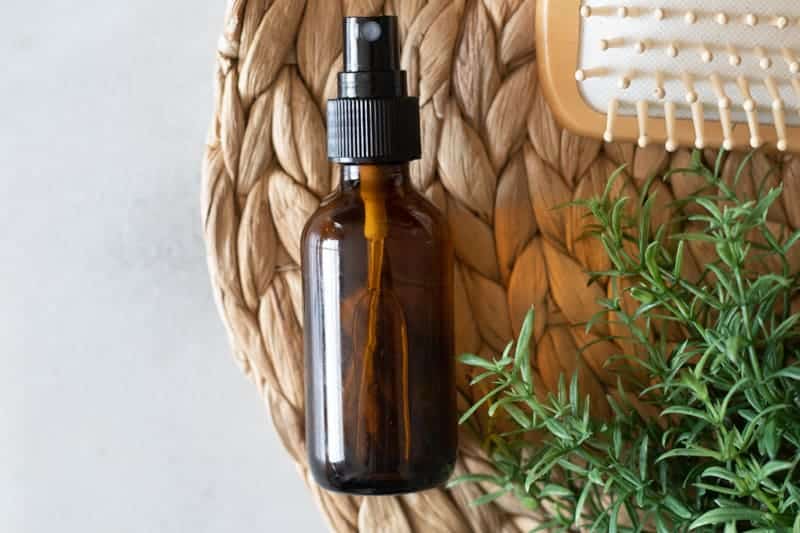 Amber spray bottle of hair spray on placemat with rosemary and tan brush.