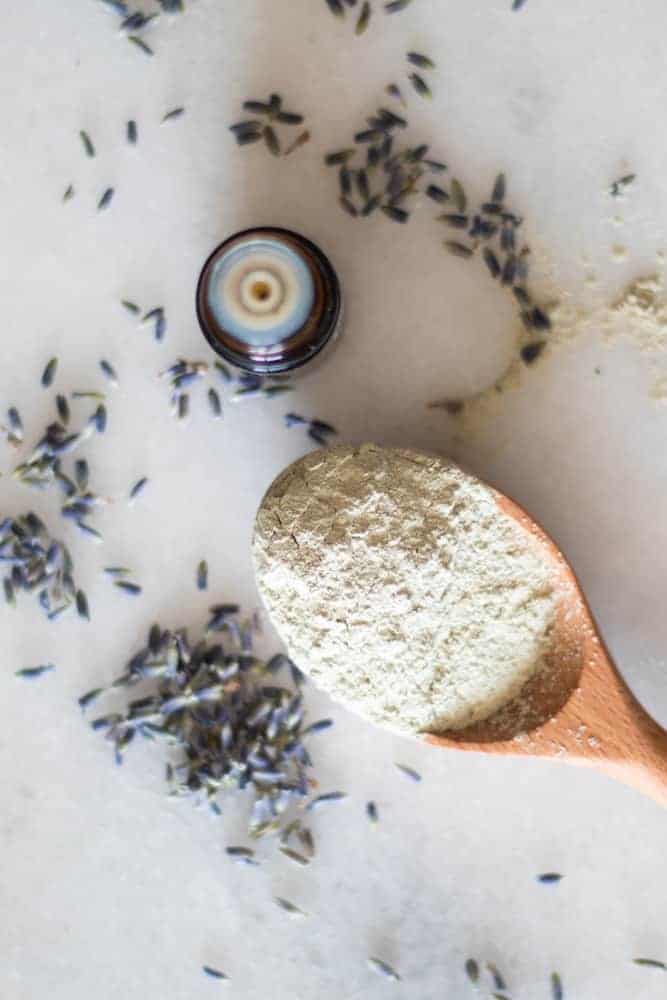 powder clay for face mask on wooden spoon with lavender springs and an essential oil bottle around it