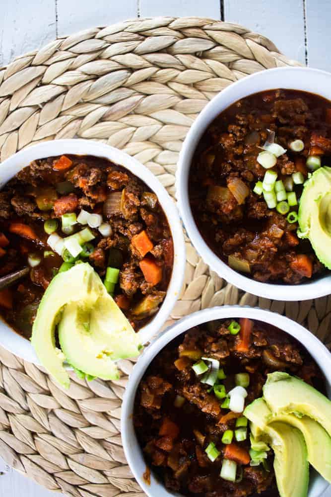 vegetable beef chili in white bowls topped with avocados on woven pad