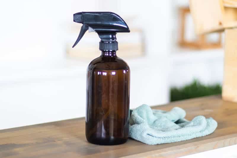 amber glass spray bottle of tea tree essential oil cleaner on wooden end table next to blue cloth in front of white wall.