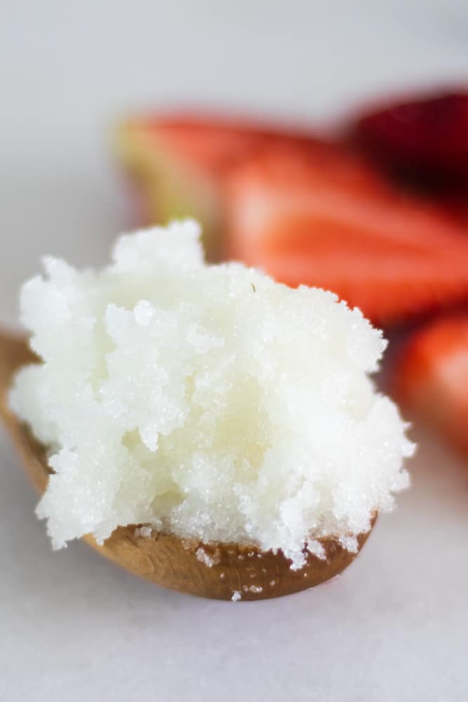 White exfoliating body scrub scoop on a spoon with sliced strawberries on white marble.