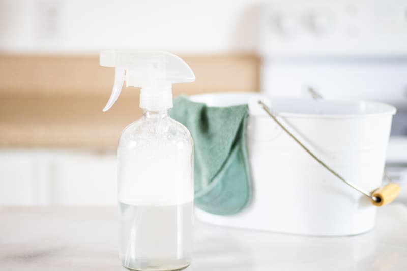 upholstery stain remover in glass spray bottle on marble table. white pail and green hand towel hanging over side.