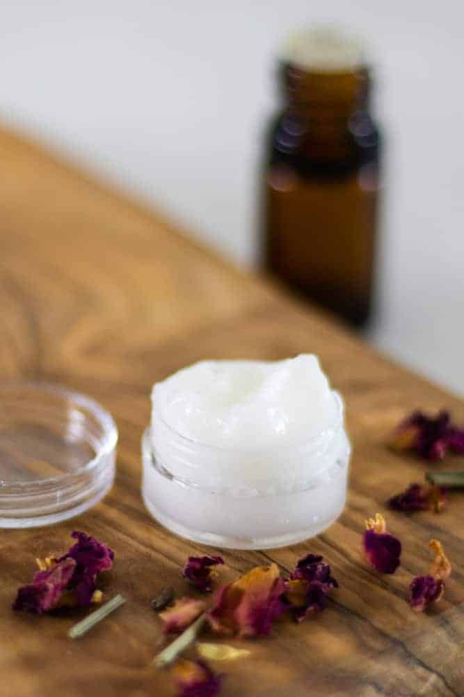 diy eye cream overflowing out of its glass tub on wooden board with essential oil bottle in background