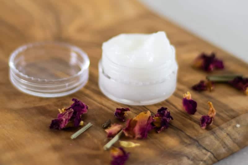 lid off of coconut oil eye cream in small container on wooden board with rose petals around it