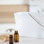 Homemade stain remover in a white wooded pail will dropper bottles and a scrubbing brush on white marble table.