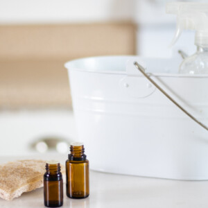 Homemade stain remover in a white wooded pail will dropper bottles and a scrubbing brush on white marble table.