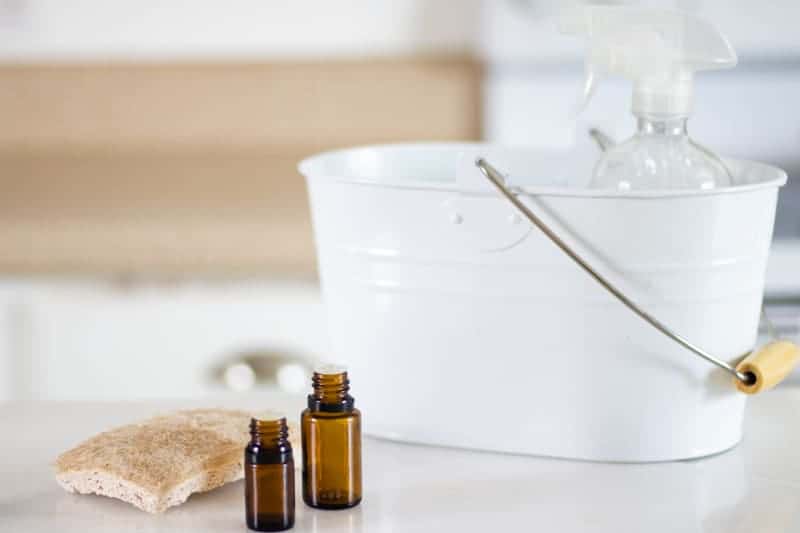 Homemade stain remover and essential oil bottles, tan sponge in white bucket on white marble table.