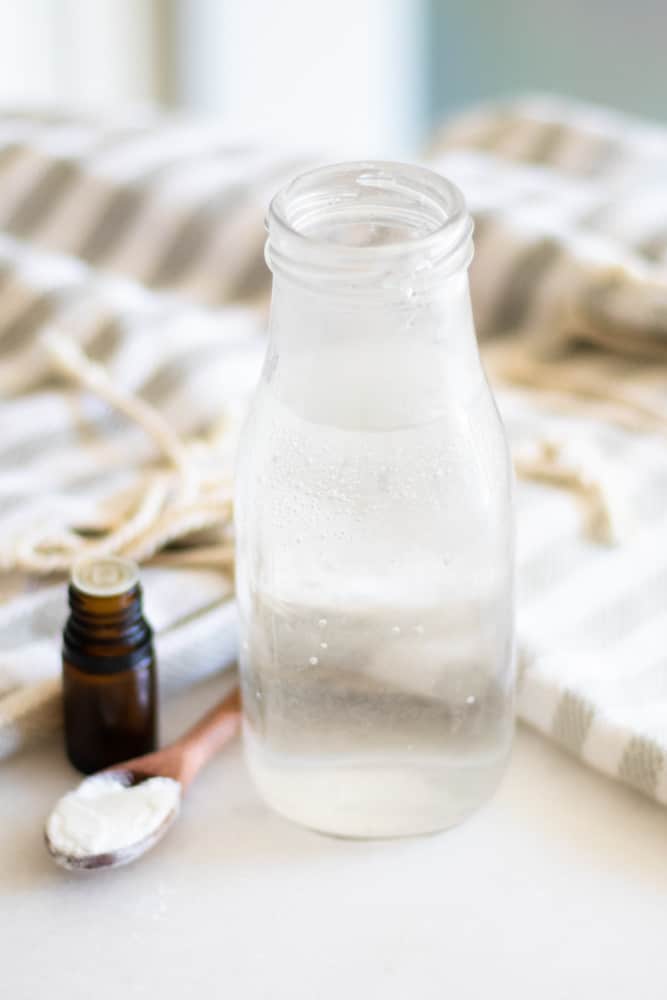 Homemade mouthwash in long neck glass container on white marble with wooden measuring spoon and essential oil bottle.