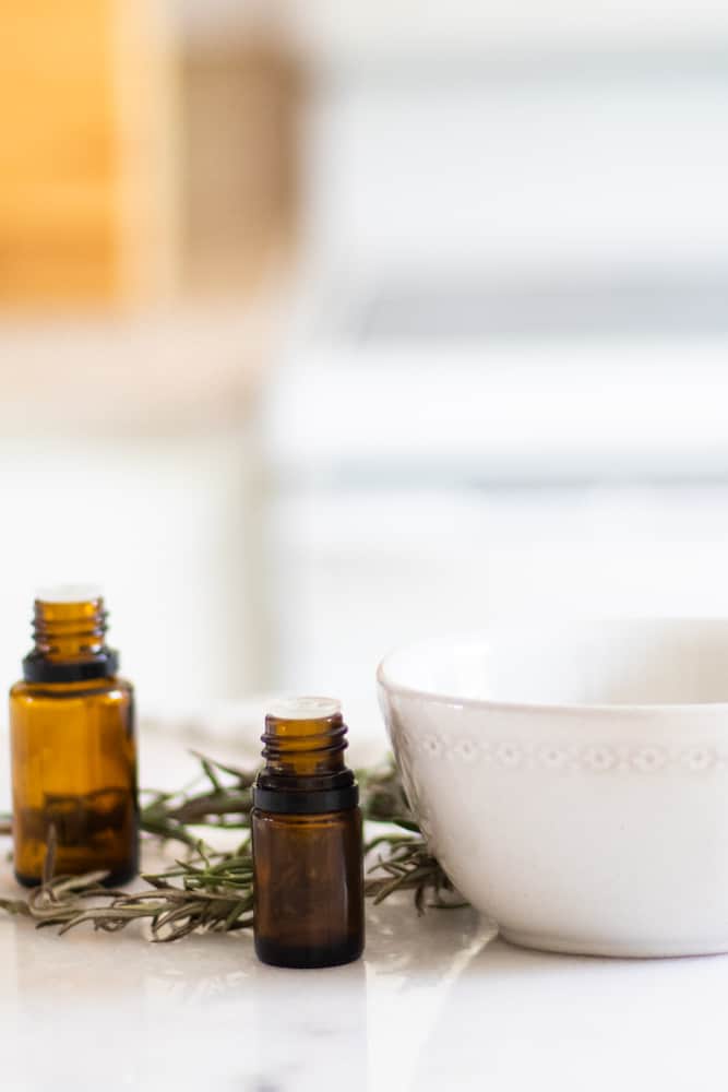 Diy deep conditioner ingredients with amber colored essential oil bottles, white bowl, and fresh rosemary on white marble.