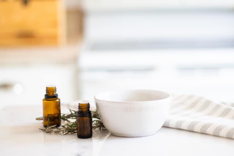 Homemade deep conditioner treatment in small white bowl on white table with amber oil bottles rosemary sprigs and a white and grey striped towel 