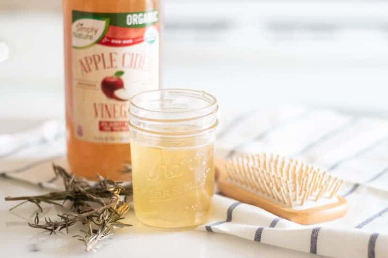 A glass mason jar of apple cider vinegar hair rinse with dried herbs and a soft bristle comb.
