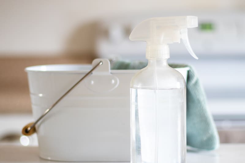 homemade mildew remover in clear glass spray bottle, with white pail and cleaning cloth.